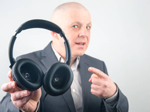 image | Bose Headphones Repair Man in a suit holds up black over-ear headphones toward the camera while pointing, as if offering them or giving a demo.